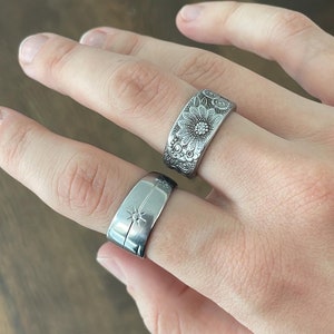 Puede incluir: Dos anillos de plata en una mano. Un anillo tiene un diseño floral y el otro tiene un diseño de estrella.