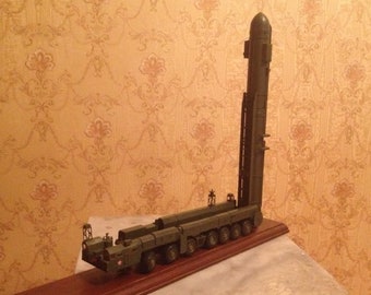 Könnte beinhalten: Ein Modell einer großen, grünen Militärrakete auf einem Transport- und Abschussfahrzeug. Die Rakete ist vertikal auf dem Fahrzeug montiert, das sechs Räder auf jeder Seite hat. Das Modell wird auf einem Holzsockel präsentiert.