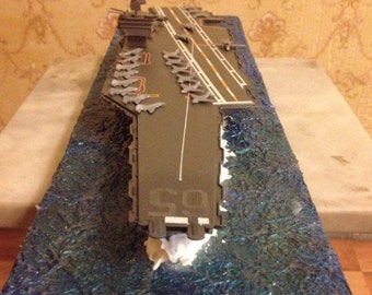 May include: A model of an aircraft carrier with a gray deck and a blue ocean base. The carrier has a number of small, gray aircraft lined up on the deck. The carrier is labeled "50".