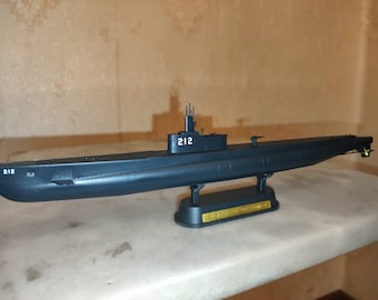 May include: A dark gray model of a submarine with the number "212" printed on the side. The submarine is on a display stand with a gold plaque.