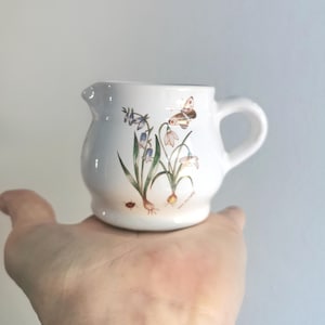 May include: White ceramic creamer with a floral design featuring blue and white flowers, green leaves, and a brown butterfly. The creamer has a rounded body and a handle.