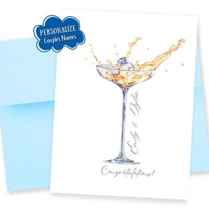 May include: A white greeting card with a champagne glass illustration. The glass contains a ring and champagne, with the words "Emily & Dylan" and "Congratulations!" written on the card. A light blue envelope is behind the card.