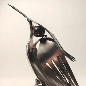 May include: A handcrafted metal bird sculpture made from repurposed silverware. The bird features a fork as a beak, spoons for the body, and a dark, round bead for an eye. The sculpture is a unique piece of art.