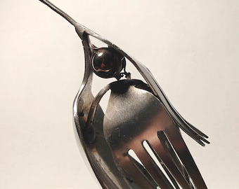 May include: A bird sculpture crafted from silver-toned metal cutlery, featuring a long pointed beak, a dark spherical eye, and a body formed from the tines and handle of a fork. The metallic surface reflects light, highlighting the curved shapes.