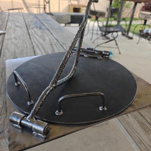 May include: A circular black metal plate featuring a hinged rebar handle assembly attached to a square base plate. The assembly includes two additional small U-shaped metal handles welded onto the surface of the circular plate, all constructed from dark, industrial-grade steel with visible weld points.