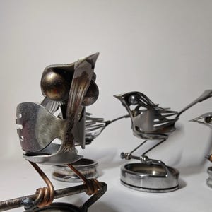 May include: Three bird sculptures crafted from repurposed silver-toned metal cutlery. Each bird features rounded metal eyes, fork-tine wings, and a tail, perched on a circular metal base with copper-colored wire accents on the legs and feet.