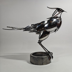 May include: A sculpture of a bird constructed from polished silver-colored metal cutlery, including spoons and forks. The bird stands on a circular, dark, weathered metal base, with its head tilted upward and its tail feathers extended horizontally.