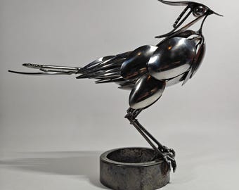 May include: A sculpture of a bird constructed from polished silver-colored metal cutlery, including spoons and forks. The bird stands on a circular, dark, weathered metal base, with its head tilted upward and its tail feathers extended horizontally.