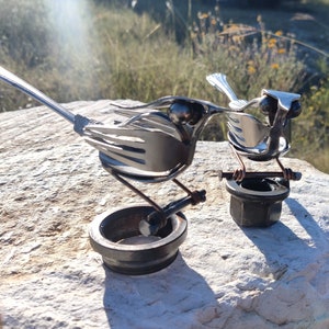 May include: Two metal bird sculptures made from recycled silverware, perched on metal bases. The birds are silver and have detailed wings and beaks.