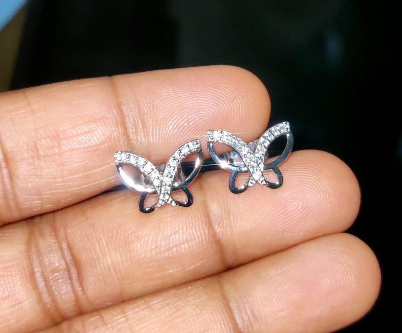 May include: A pair of silver butterfly stud earrings. Each earring has a butterfly design with clear, sparkling stones set along the wings. The earrings are displayed on a hand against a dark background.