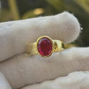 May include: A gold ring featuring an oval, deep red gemstone. The ring has a simple, polished band and a bezel setting that securely holds the gemstone. The ring is displayed on a white surface.