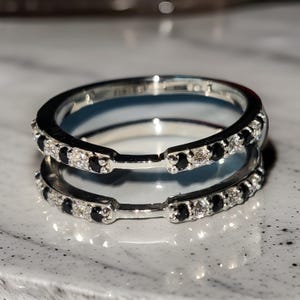 May include: A silver ring guard with a double band design. Each band is adorned with alternating black and clear gemstones. The ring is set against a light-colored, speckled surface, creating a contrast that highlights the ring's details.