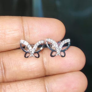 May include: A pair of butterfly stud earrings. The earrings are silver-toned with a black outline and are encrusted with small, sparkling stones. The butterfly wings are open, and the design is delicate and detailed.