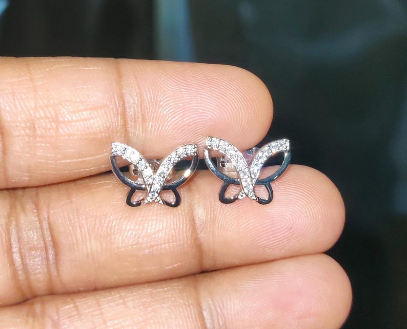 May include: A pair of butterfly stud earrings. The earrings are silver-toned with a black outline and are encrusted with small, sparkling stones. The butterfly wings are open, and the design is delicate and detailed.