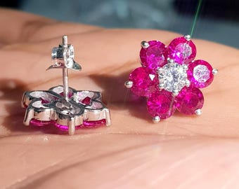 May include: A pair of flower-shaped stud earrings. Each earring features a cluster of six magenta-colored gemstones surrounding a central, clear gemstone. The earrings are set in a silver-colored metal.