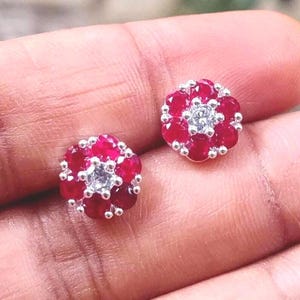 May include: A pair of stud earrings featuring a floral design. Each earring has a central cluster of small, clear stones surrounded by a ring of vibrant red gemstones. The earrings are set in a silver-colored metal.