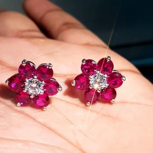 Peut inclure: Une paire de boucles d'oreilles &agrave; clous en forme de fleur. Chaque boucle d'oreille pr&eacute;sente un groupe de pierres pr&eacute;cieuses rose fonc&eacute; dispos&eacute;es autour d'une pierre pr&eacute;cieuse centrale, claire et ronde. Les boucles d'oreilles ont un sertissage en m&eacute;tal argent&eacute;.