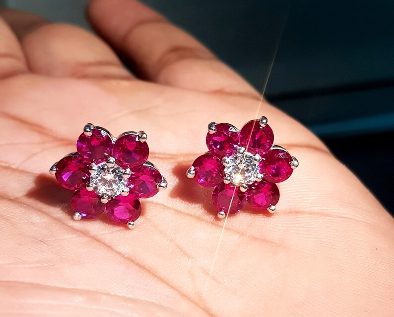 Peut inclure: Une paire de boucles d'oreilles &agrave; clous en forme de fleur. Chaque boucle d'oreille pr&eacute;sente un groupe de pierres pr&eacute;cieuses rose fonc&eacute; dispos&eacute;es autour d'une pierre pr&eacute;cieuse centrale, claire et ronde. Les boucles d'oreilles ont un sertissage en m&eacute;tal argent&eacute;.