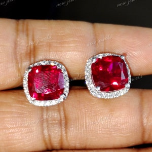 May include: A pair of square-cut ruby stud earrings with a halo of small, clear gemstones. The rubies are a deep red color, and the surrounding stones are set in a silver or white metal. The earrings are displayed against a neutral background.