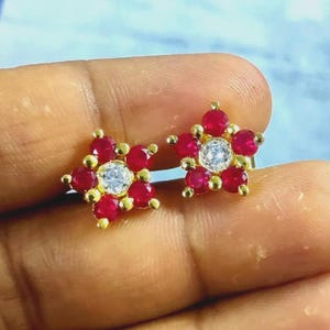 May include: A pair of gold-toned flower-shaped stud earrings. Each earring features a central, clear, round gemstone surrounded by red gemstones. The earrings are set against a neutral background.
