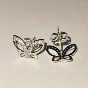 May include: A pair of silver butterfly stud earrings. One earring is in focus, showcasing a detailed design with small, clear stones. The other earring is out of focus, showing the post and butterfly shape.