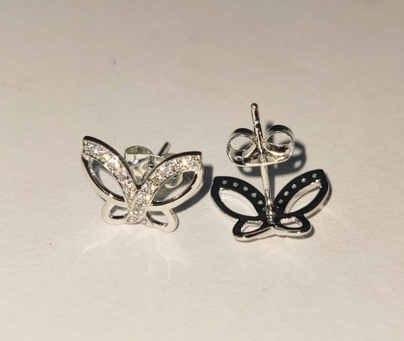 May include: A pair of silver butterfly stud earrings. One earring is in focus, showcasing a detailed design with small, clear stones. The other earring is out of focus, showing the post and butterfly shape.