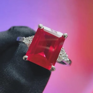 May include: A silver ring featuring a large, rectangular, bright red gemstone as the focal point. The ring has smaller, clear stones on either side of the main stone, adding sparkle. The ring is set against a colorful, blurred background.