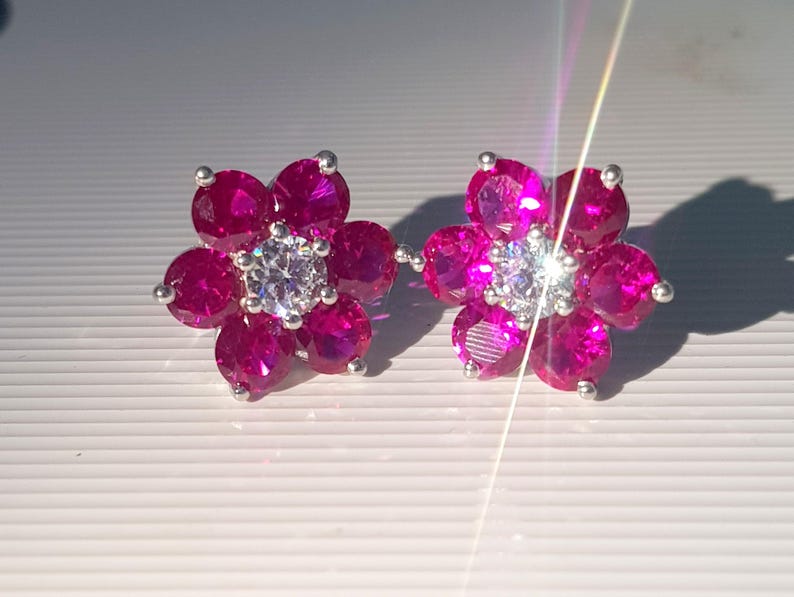 Peut inclure: Une paire de boucles d'oreilles &agrave; clous en forme de fleur. Chaque boucle d'oreille pr&eacute;sente un groupe de pierres pr&eacute;cieuses magenta vif dispos&eacute;es autour d'une pierre centrale claire et &eacute;tincelante. Les boucles d'oreilles sont serties dans un m&eacute;tal argent&eacute;.