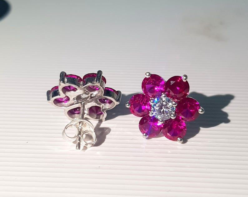 Peut inclure: Une paire de boucles d'oreilles &agrave; clous en forme de fleur. Chaque boucle d'oreille pr&eacute;sente un sertissage en m&eacute;tal argent&eacute; avec six pierres pr&eacute;cieuses magenta dispos&eacute;es en forme de p&eacute;tales. Un diamant clair et rond est au centre de chaque fleur.