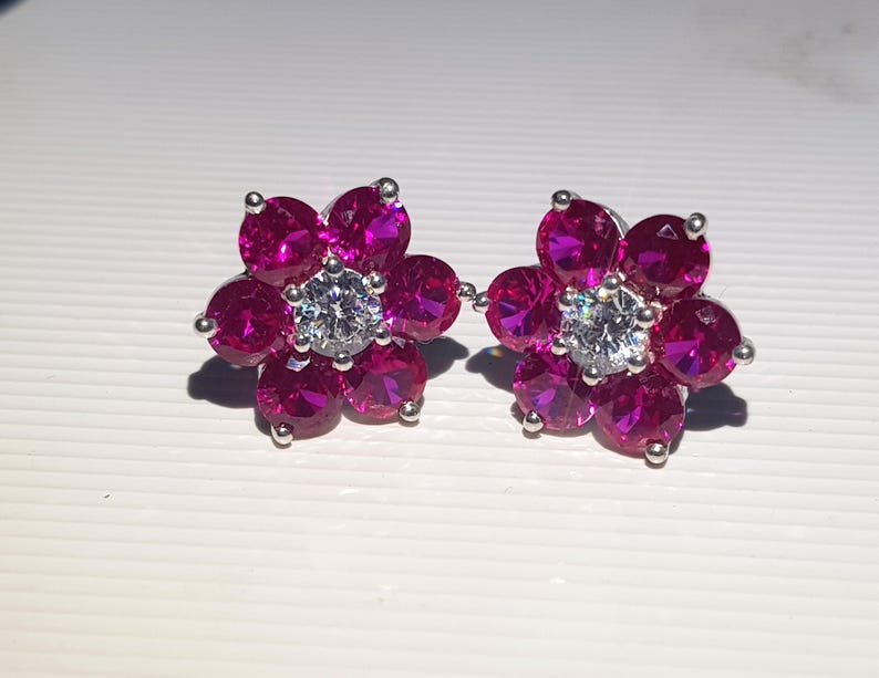 Peut inclure: Une paire de boucles d'oreilles &agrave; clous en forme de fleur. Chaque boucle d'oreille pr&eacute;sente six pierres pr&eacute;cieuses magenta entourant une pierre centrale transparente. Les boucles d'oreilles sont serties dans un m&eacute;tal argent&eacute;.