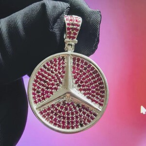 May include: A silver pendant featuring a Mercedes-Benz logo design. The logo's star and the surrounding circle are encrusted with small, red gemstones. The pendant is suspended from a silver bail.