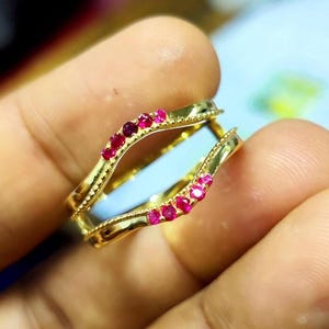 May include: A gold-colored ring with a double band design. The ring is adorned with small, round, red gemstones. The ring is held between fingers, showcasing its intricate details and craftsmanship. The ring's design is elegant and eye-catching.