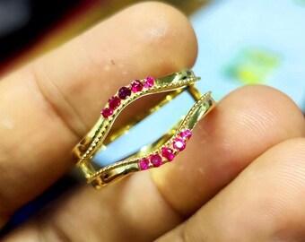 May include: A gold-colored ring with a double band design. The ring is adorned with small, round, red gemstones. The ring is held between fingers, showcasing its intricate details and craftsmanship. The ring's design is elegant and eye-catching.