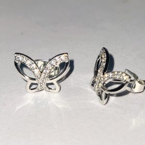 May include: A pair of silver butterfly stud earrings. Each earring has a butterfly design with a row of small, clear gemstones along the wings. The earrings are displayed on a white surface.