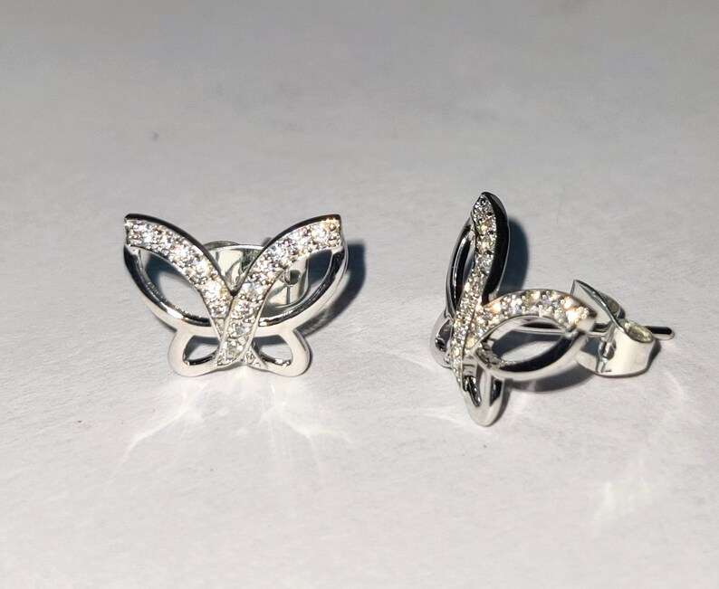 May include: A pair of silver butterfly stud earrings. Each earring has a butterfly design with a row of small, clear gemstones along the wings. The earrings are displayed on a white surface.