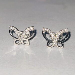 May include: A pair of silver butterfly stud earrings. Each earring has a butterfly design with clear, sparkling stones in the wings and a polished silver frame. The earrings are on a light grey surface.