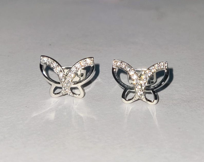 May include: A pair of silver butterfly stud earrings. Each earring has a butterfly design with clear, sparkling stones in the wings and a polished silver frame. The earrings are on a light grey surface.