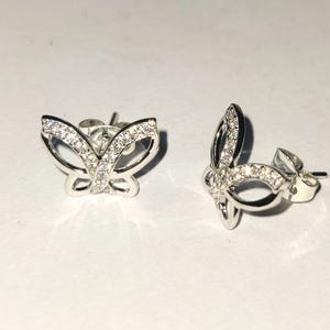 May include: A pair of silver butterfly stud earrings. Each earring has a butterfly design with a clear gemstone-studded wing outline. The earrings are presented on a white surface, highlighting their delicate details.