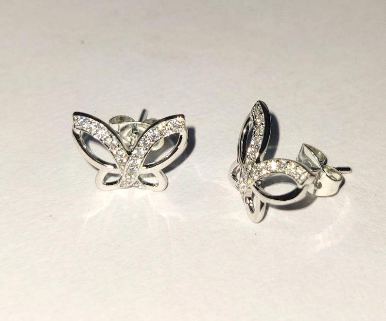May include: A pair of silver butterfly stud earrings. Each earring has a butterfly design with a clear gemstone-studded wing outline. The earrings are presented on a white surface, highlighting their delicate details.