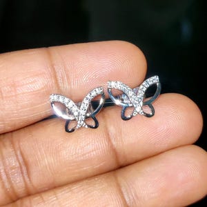 May include: A pair of silver butterfly stud earrings. Each earring has a butterfly design with clear, sparkling stones set along the wings. The earrings are displayed against a dark background.