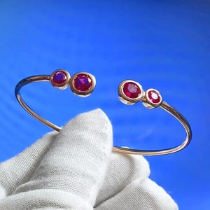 May include: A gold-toned open cuff bracelet with three round, vibrant red gemstones on each end. The bracelet is held against a solid blue background, showcasing the jewelry's design and color.