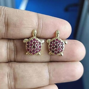 May include: A pair of gold-colored turtle stud earrings. The turtle shells are encrusted with small, round, ruby-colored stones. The earrings have a polished finish and are designed with detailed turtle features.