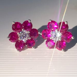 Peut inclure: Une paire de boucles d'oreilles &agrave; clous en forme de fleur. Chaque boucle d'oreille pr&eacute;sente un groupe de pierres pr&eacute;cieuses magenta vif dispos&eacute;es autour d'une pierre centrale blanche &eacute;tincelante. Les boucles d'oreilles ont un sertissage en m&eacute;tal argent&eacute;.