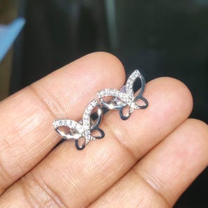 May include: A pair of silver butterfly stud earrings. Each earring features a butterfly design with clear, sparkling stones set along the wings. The earrings are displayed against a neutral background.