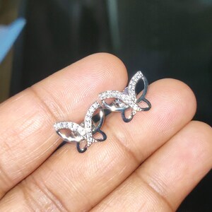 May include: A pair of silver butterfly stud earrings. Each earring has a butterfly design with a clear, sparkling stone-studded wing outline. The earrings are shown against a neutral background.