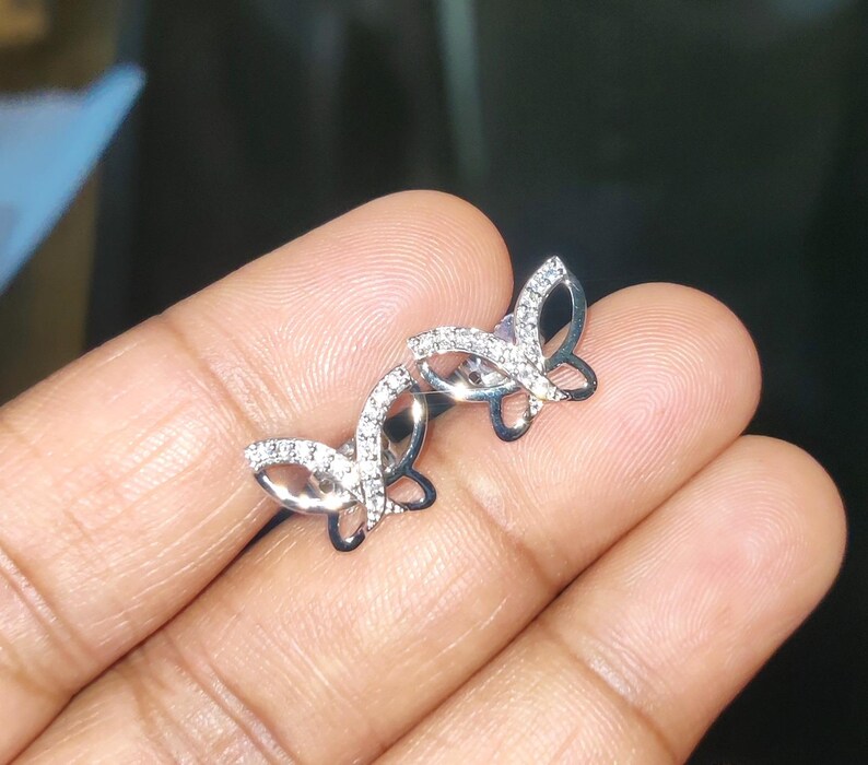 May include: A pair of silver butterfly stud earrings. Each earring has a butterfly design with a clear, sparkling stone-studded wing outline. The earrings are shown against a neutral background.