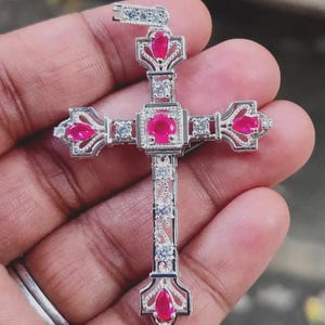 May include: A silver pendant in the shape of a cross, adorned with vibrant red gemstones and clear accent stones. The cross features an ornate design with a bail for a chain. The gemstones are set in a detailed, filigree-style metalwork.