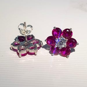 Peut inclure: Une paire de boucles d'oreilles &agrave; clous en forme de fleur. Chaque boucle d'oreille pr&eacute;sente un groupe de pierres pr&eacute;cieuses rose fonc&eacute; dispos&eacute;es autour d'une pierre pr&eacute;cieuse centrale et transparente. Les boucles d'oreilles ont un sertissage et un fermoir en m&eacute;tal argent&eacute;.
