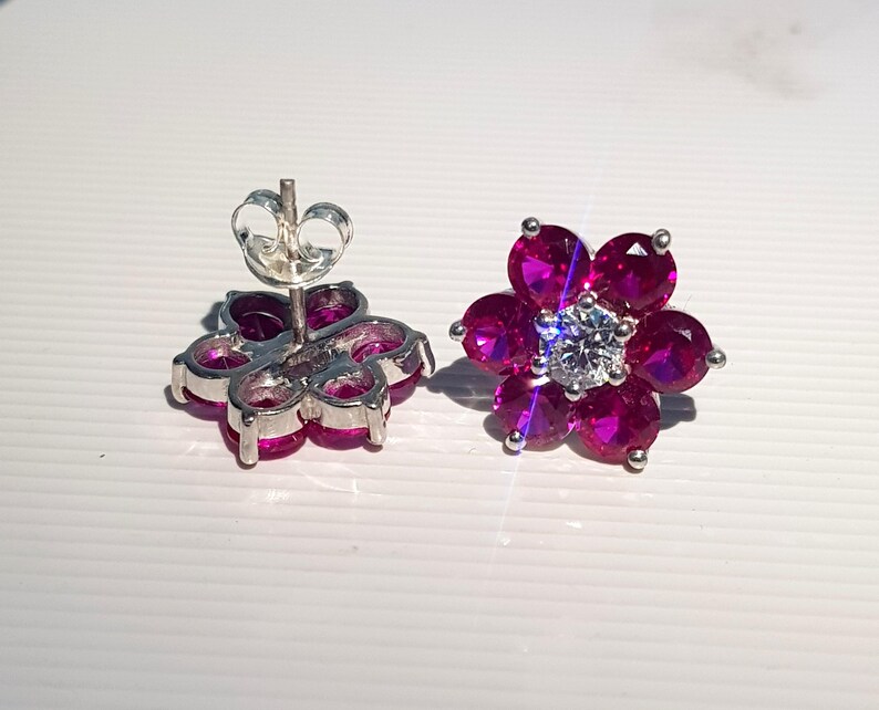 Peut inclure: Une paire de boucles d'oreilles &agrave; clous en forme de fleur. Chaque boucle d'oreille pr&eacute;sente un groupe de pierres pr&eacute;cieuses rose fonc&eacute; dispos&eacute;es autour d'une pierre pr&eacute;cieuse centrale et transparente. Les boucles d'oreilles ont un sertissage et un fermoir en m&eacute;tal argent&eacute;.