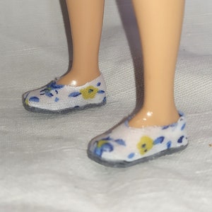 May include: A pair of miniature doll shoes. The shoes are white with a floral pattern in yellow and blue. The soles are gray. The shoes are on a doll's feet.